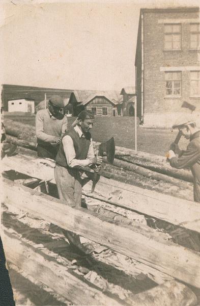 1956-1.jpg - Ciosanie drzewa na dom Zbigniewa Wierdaka. Od lewej Henryk Fruga, Władysław Kołacz /z toporem/ i Stanisław Kołacz. W głębi szkoła - jeszcze bez skrzydła północnego oraz "surowy" Dom Ludowy